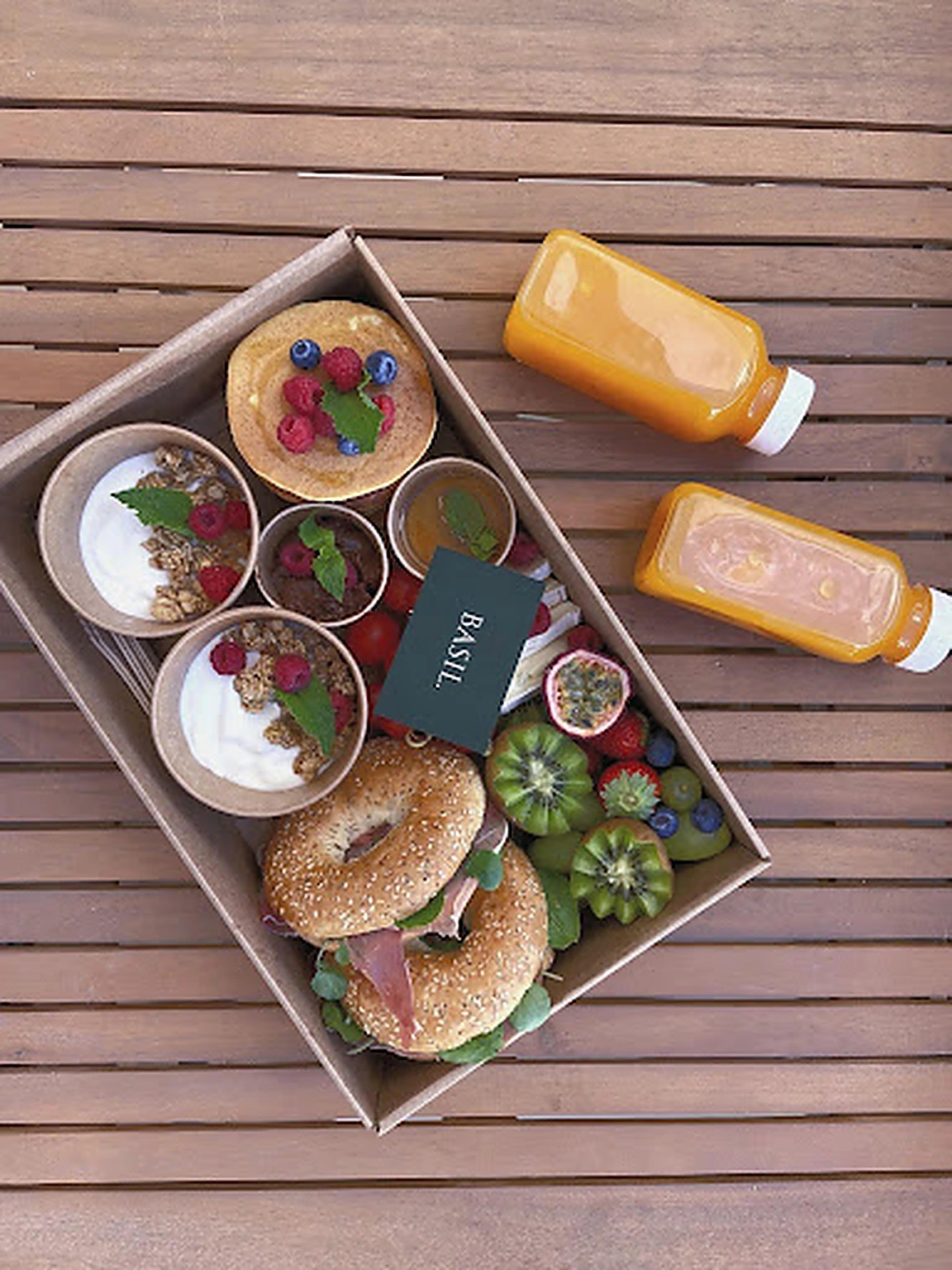 A Basil food box with bagels, pancakes, yoghurt, fresh fruit and juices on a wooden terrace table
