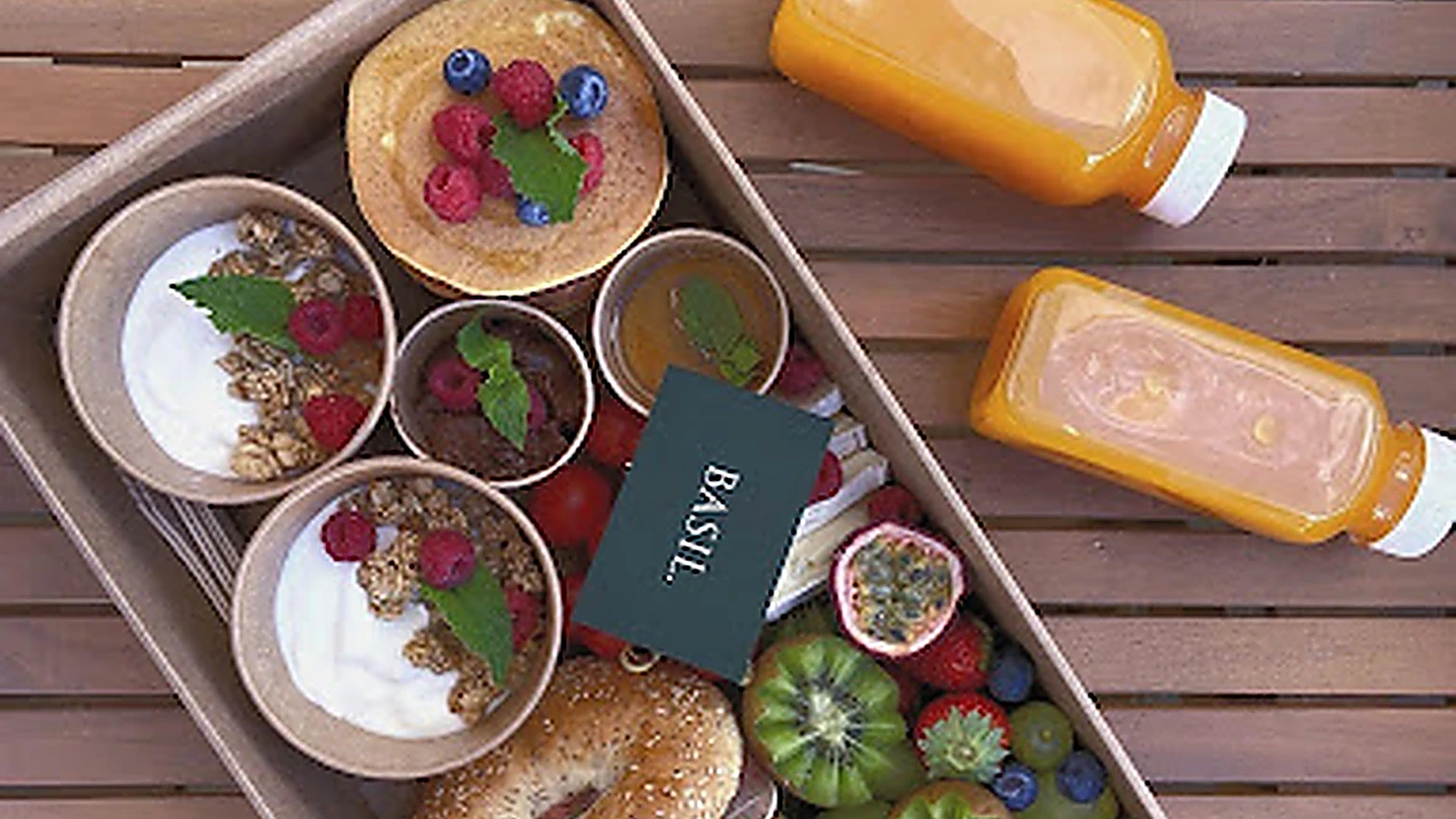 A Basil food box with bagels, pancakes, yoghurt, fresh fruit and juices on a wooden terrace table