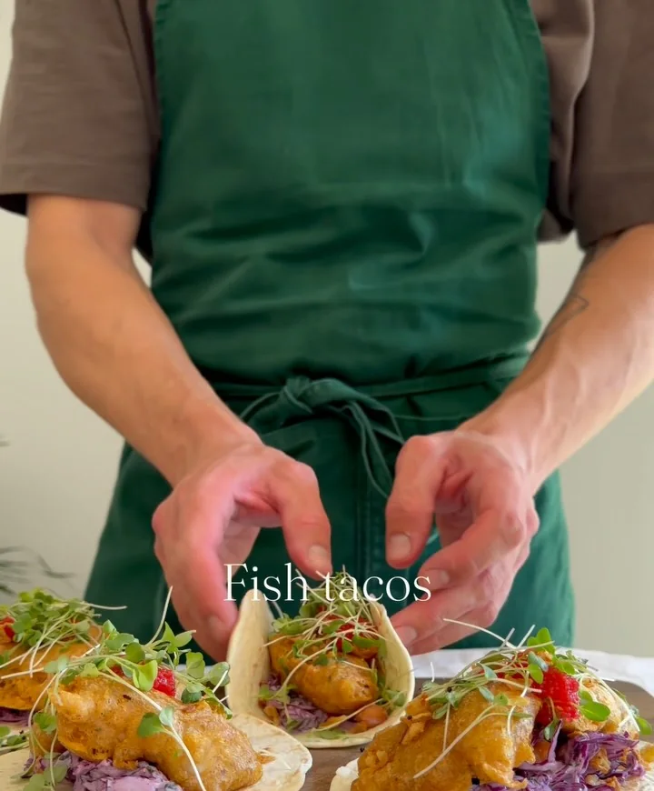 A chef in an olive apron finishing sea bass tacos on a wooden board