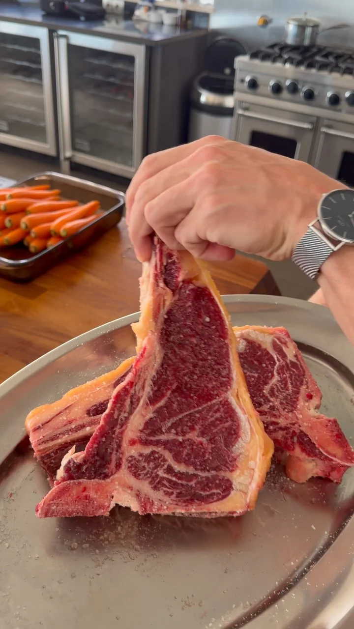 A chef's hand lifting two dry-aged tomahawk steaks from a steel tray in a villa kitchen